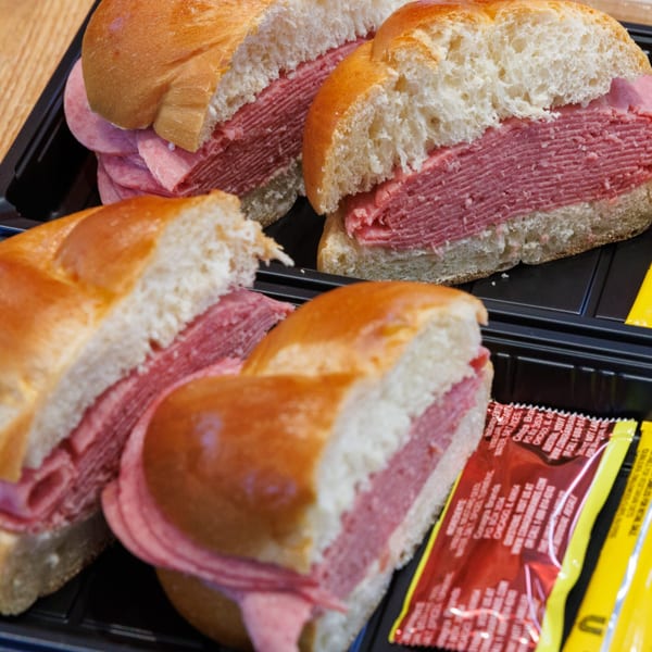 Four deli sandwiches with thick layers of meat on challah buns, served in black plastic containers.