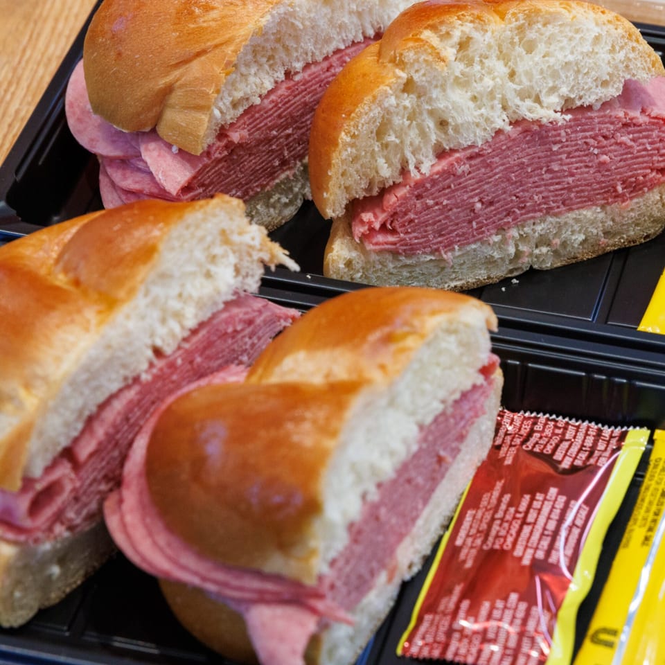 Four deli sandwiches with thick layers of meat on challah buns, served in black plastic containers.