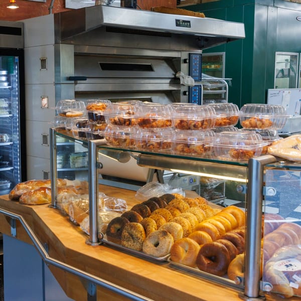 A deli counter with a variety of fresh bagels, pastries, and bread, with a large oven in the background.