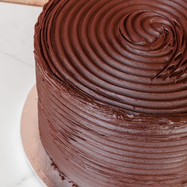 Close-up of a rich, dark chocolate cake with swirled frosting on a white marble surface.