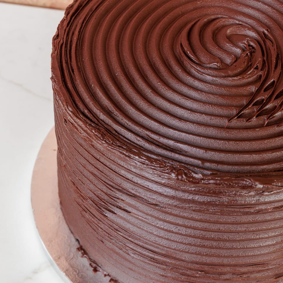 Close-up of a rich, dark chocolate cake with swirled frosting on a white marble surface.