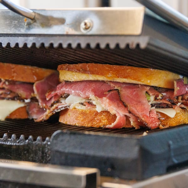 Two Wolfie's deli sandwiches with pastrami, cheese, and sauerkraut being pressed on a grill.